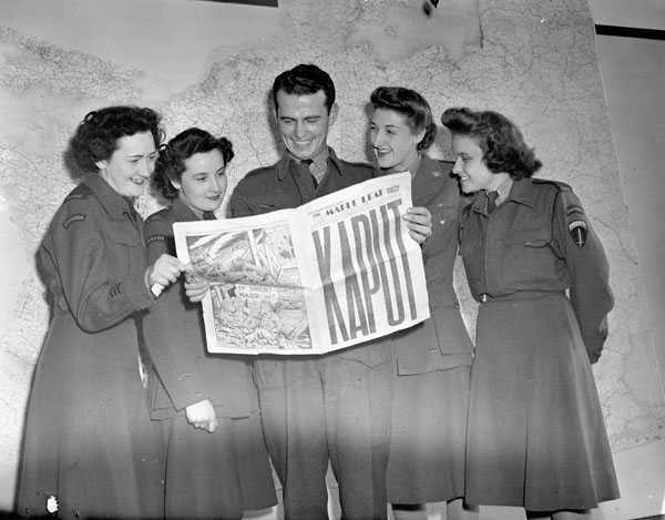 Photographie en noir et blanc – Un homme tient un journal ouvert à une page centrale; la une est couverte de grosses lettres indiquant « KAPUT ». Quatre femmes l’entourent, lisant le journal avec lui.